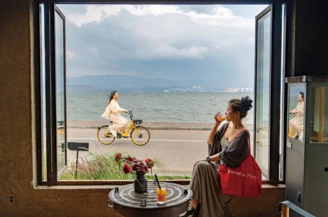 ▲2025年4月23日，一名来自安徽的游客在大理洱海生态廊道咖啡馆内欣赏洱海风光，一名骑行的游客从窗前经过 图/新华社