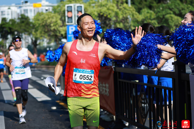 2025年12月21日，广州马拉松的赛道旁，一位跑者正笑容满面地与啦啦队员互动。阳光明媚的赛道上，既展现了马拉松运动的快乐氛围，也传递出这场城市赛事里的温暖联结。