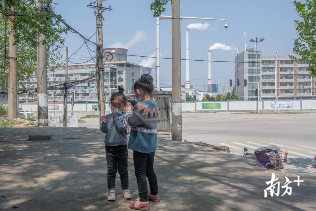 4月12日，武钢炼钢厂外，一个小女孩正帮助自己的妹妹戴好口罩，其不远处的烟囱正向外排着浓烟，武汉大大小小的企业、工厂正有序复工复产。